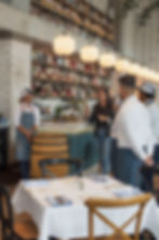 In a stylish bar-restaurant, a woman briefs staff in white shirts and aprons beneath pendant lights and liquor shelves.