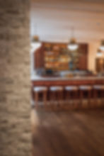 Stylish bar interior with wooden barstools, a stocked liquor shelf, and glowing pendant lights. A vase with flowers adds decor. Quiet ambiance.