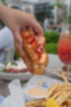 Hand holding a lobster roll on an outdoor patio, with fries and a pink drink beside blurred diners in the background.