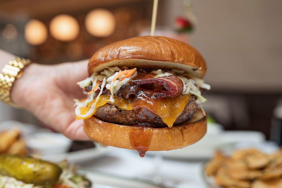 A hand holds a juicy cheeseburger with coleslaw and sauce dripping. Background shows blurred dining setting with warm lights.