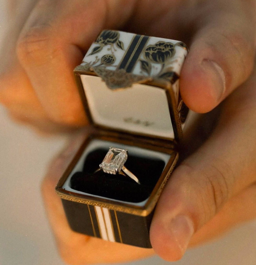 An emerald-cut diamond proposal ring resting inside a hand-painted black and gold porcelain peony ring box, held open on a beach in warm sunset light.
