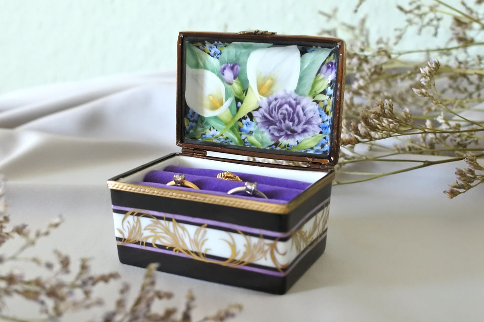 An open, bespoke porcelain ring box. The inside of the lid features hand-painted white calla lilies, a purple carnation, and blue forget-me-nots. Below, vintage diamond rings rest securely in vibrant purple velvet slots.