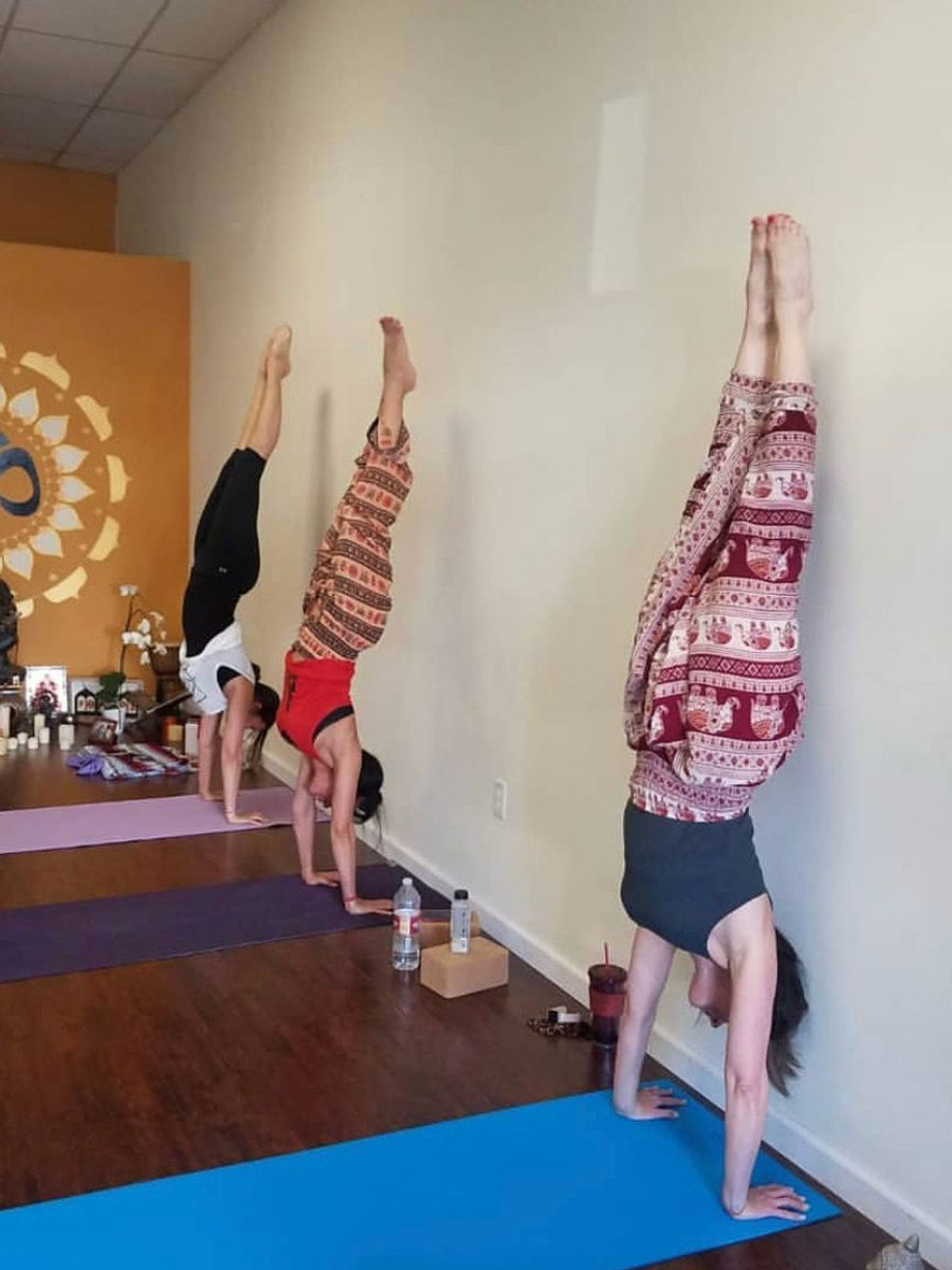 Three students holding handstands against the studio wall, a mandala decal between them and blocks on the floor