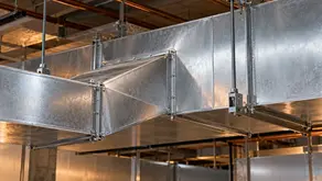 Galvanized rectangular duct transition joining a larger overhead ventilation trunk, with flanges, seams and support hangers.