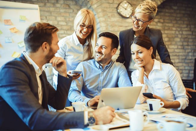 Five coworkers smile around a laptop in a brick-walled office meeting room, sharing a phone and coffee.