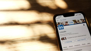 Smartphone displaying LinkedIn profile on wooden table with shadows. The screen shows LinkedIn logo and profile details. Mood is calm.