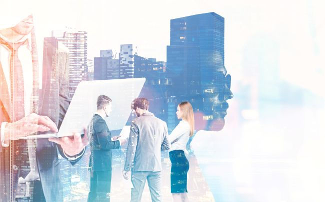 Businesspeople in suits discuss in front of a cityscape. A large, transparent figure with a laptop and skyscrapers overlay the scene.