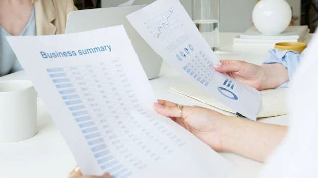 Two people holding business summary papers with charts. Office setting, laptop in background. Papers have blue graphs and text.