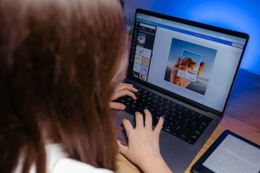 Person using a laptop, editing a photo of a dog with ocean background. Bright blue screen lighting and tablet on wooden desk.
