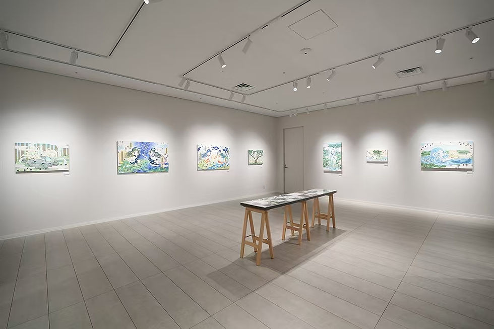 明るい白壁のギャラリーに、絵画が壁に並び、中央の作業台に作品資料が置かれた静かな展示室