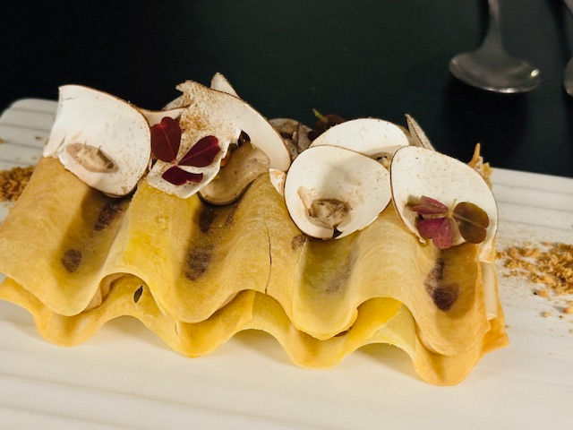 Curved pasta sheet topped with sliced mushrooms, herbs, and dusting of spices on a white plate, creating an elegant presentation.