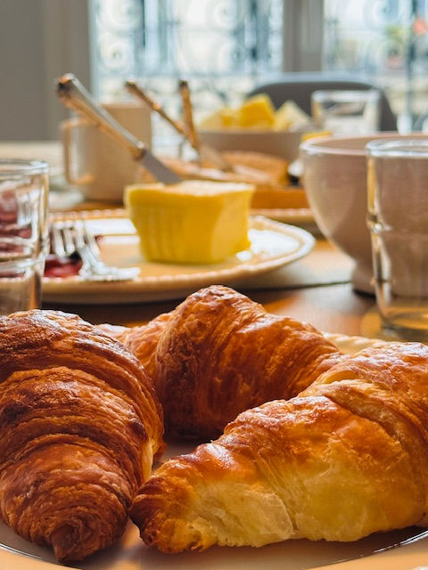 Croissants on a plate in a bright kitchen. Butter, jam, and tableware in the background. Sunlit window with ornate metalwork visible. Cozy mood.