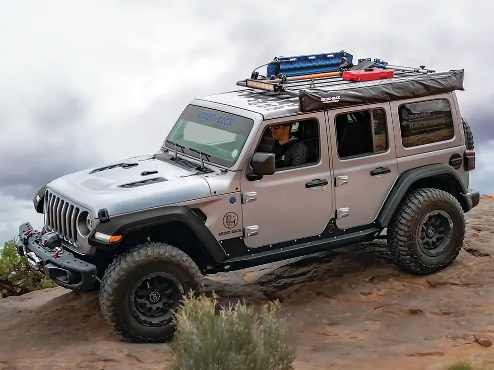 Silver Jeep with roof gear driving on rocky terrain under cloudy skies. Decals on the side. Rugged and adventurous mood.