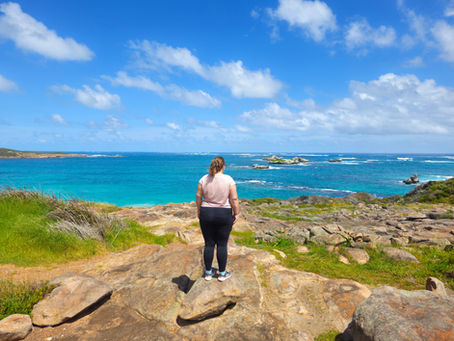 A woman standing on a rocky outcrop overlooking a wide turquoise ocean under a bright blue sky, reflecting on finding herself while navigating a period of life in limbo.