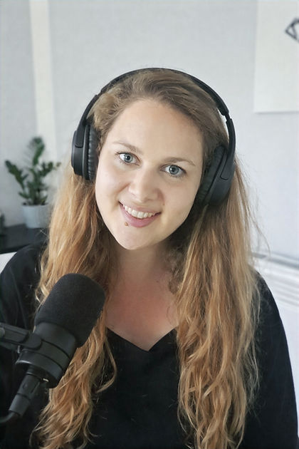 Long blonde haired lady smiling, wearing a black dress, over the head black headphones with a black microphone in front of her, with a blurry background of a home office