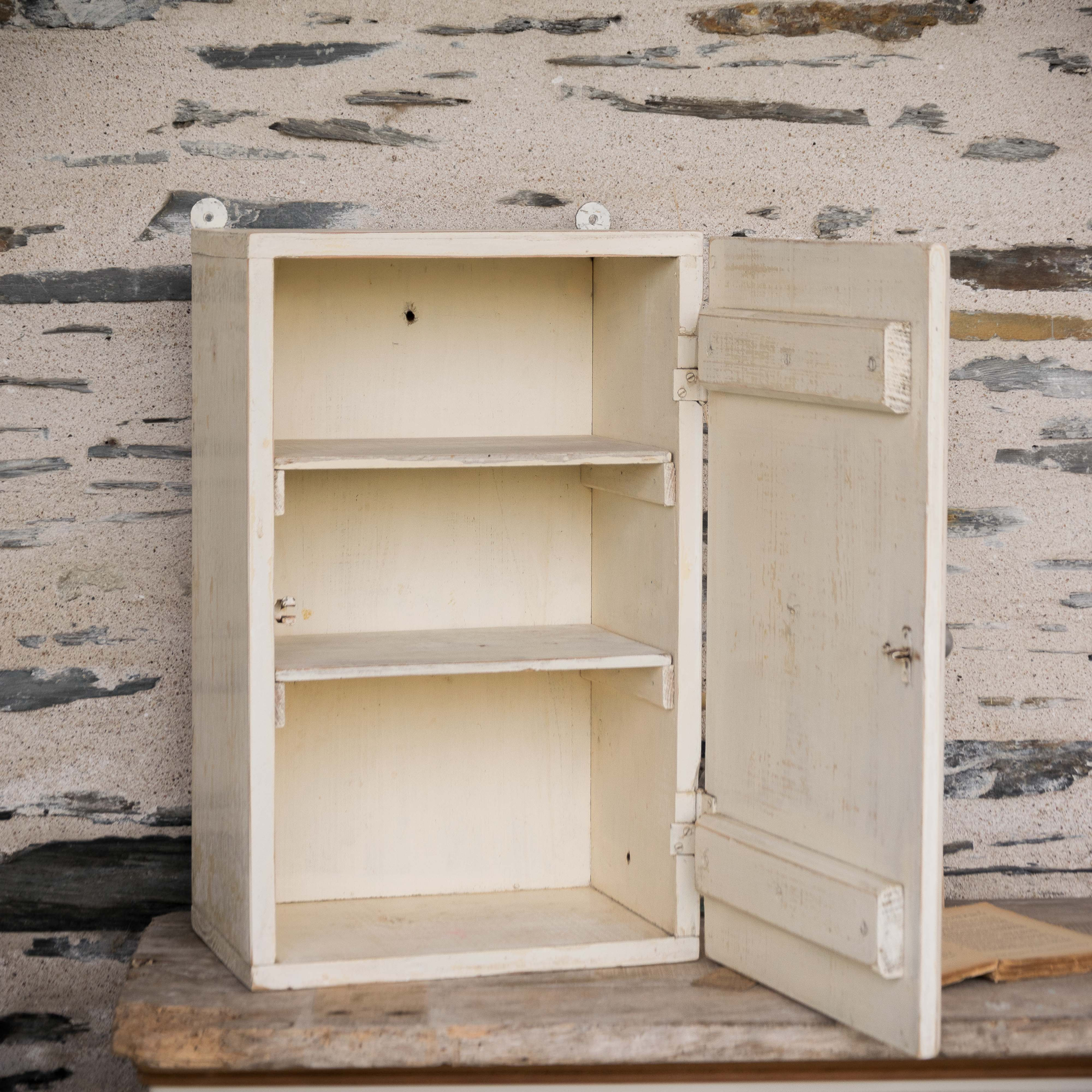 Ancienne armoire à pharmacie murale en bois patiné blanc avec croix rouge, porte ouvrante et étagères intérieures, style broc
