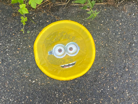 Een vergeelde gele frisbee met een vervormd Minion-gezicht ligt plat op het asfalt, omringd door onkruid. Op de voorgrond zijn de benen en loopschoenen van een vrouw zichtbaar, die er van bovenaf naar kijkt. Het beeld suggereert op ironische wijze gezichtsverlies, vergane glorie en een merk dat zijn frisheid en relevantie verloren is.