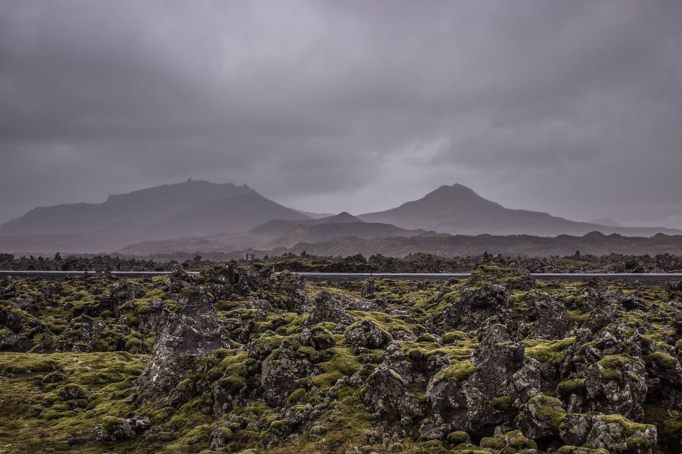 Mossy lava field beneath dark cloudy sky, with distant mountains and a road across the middle, creating a stark, moody landscape in Iceland.
