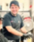 A young man wearing a black shirt, apron, hat, and gloves preparing food in a kitchen.