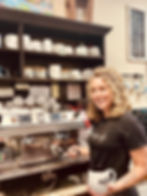 A smiling, curly-haired woman in a black shirt operating a coffee machine while holding a cup of cof