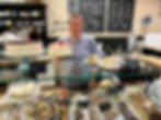 A woman wearing a pale blue shirt behind a dessert case, holding a white frosted cake topped with sl