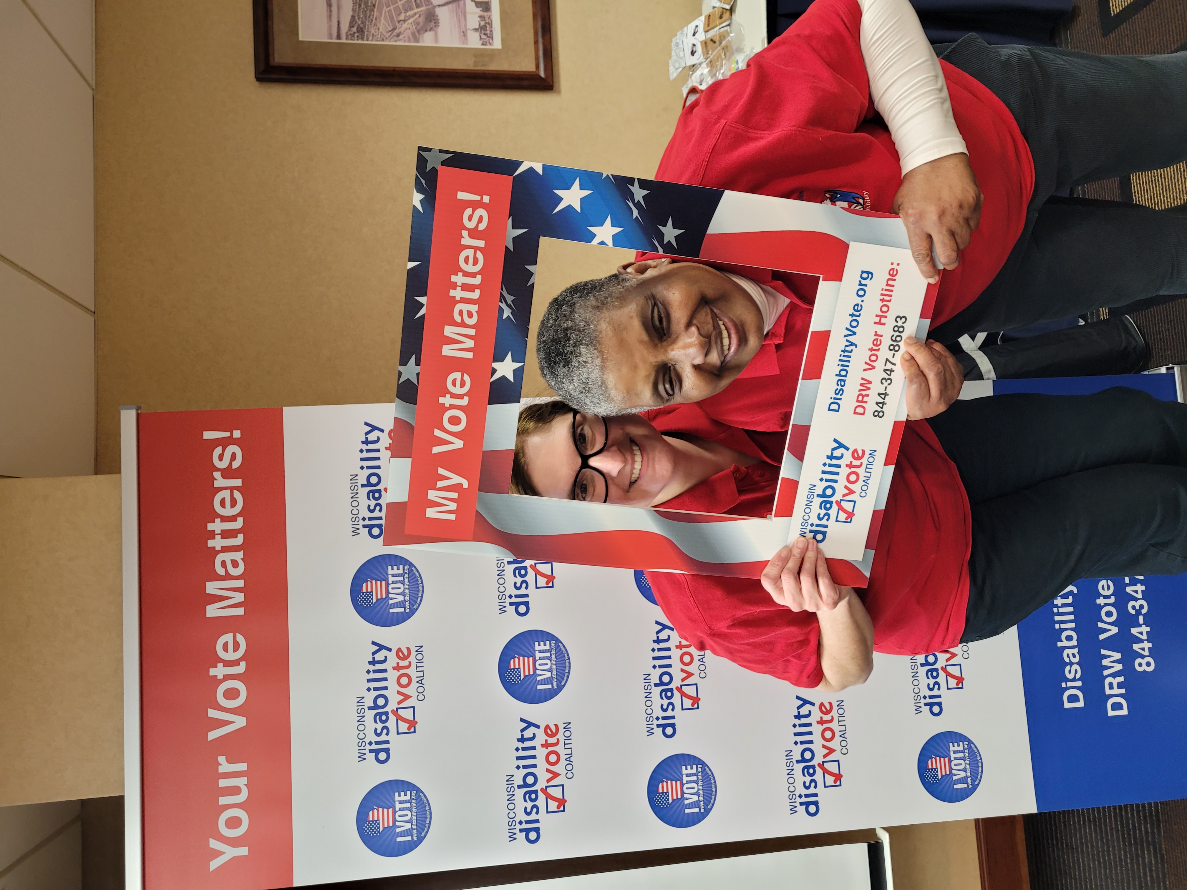 A white woman with glasses and an African American woman stand holding a picture frame with reminders to vote.