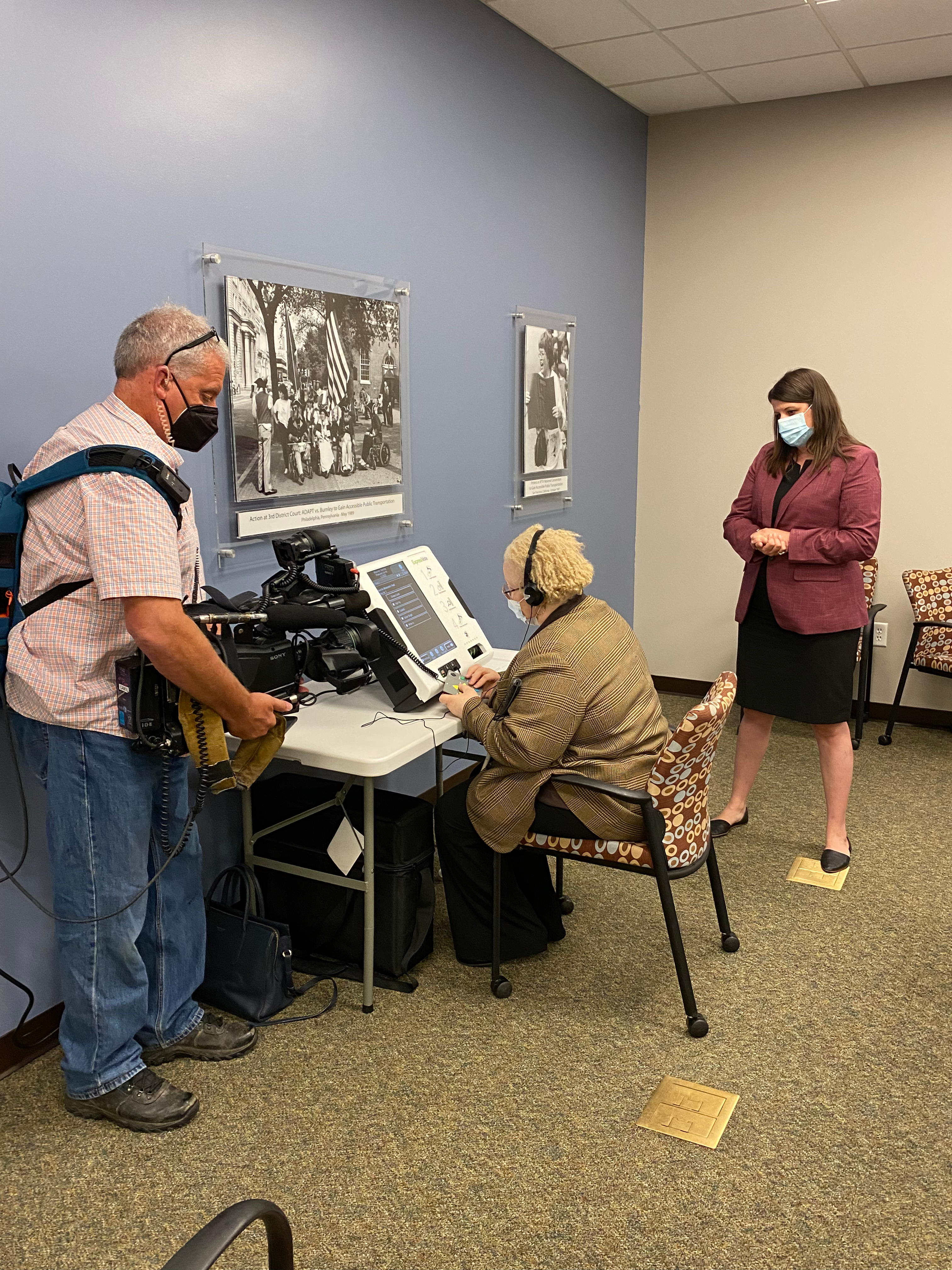 A cameraman video tapes a seated woman wearing headphones as she uses an accessible voting machine.