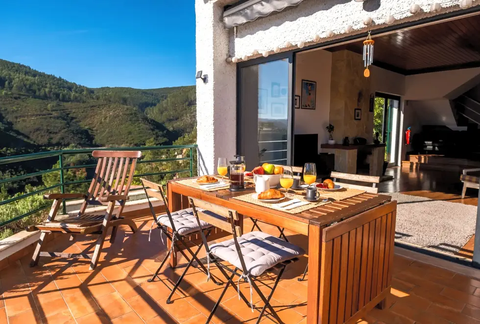 Blue Lake House’s sunny day at the balcony, large outdoor table with breakfast, sun lounger, and bright living room in the background.