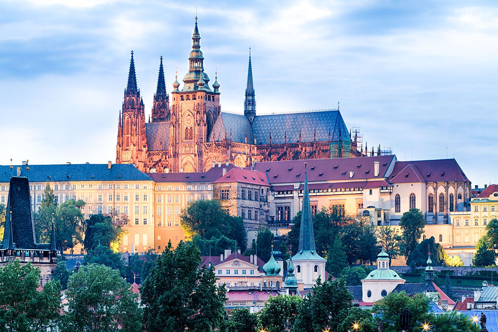 Majestic castle with tall spires under a blue sky, surrounded by lush trees. Warm evening light highlights detailed architecture.