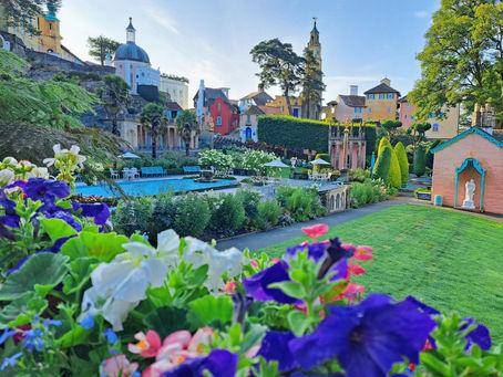 Colorful flower-filled garden with a blue pool, pastel buildings and church towers on a sunny hillside, peaceful and picturesque.