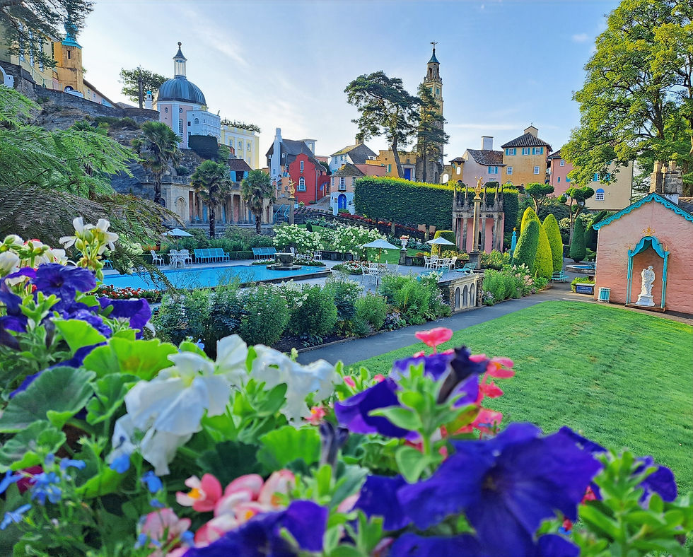 Colorful flower-filled garden with a blue pool, pastel buildings and church towers on a sunny hillside, peaceful and picturesque.