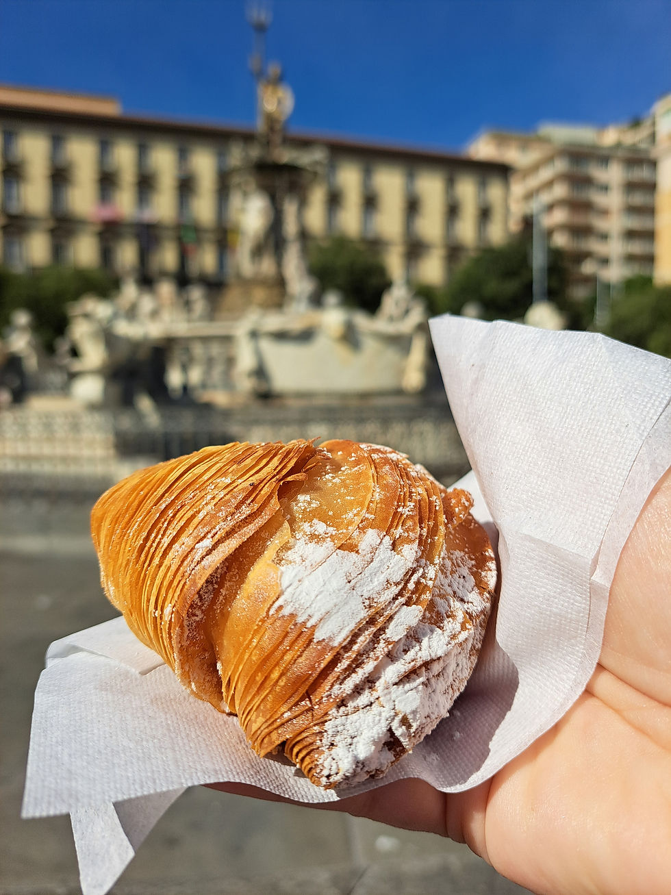 Hand holding a powdered croissant-like pastry in front of a blurred sunny fountain and buildings under a clear blue sky