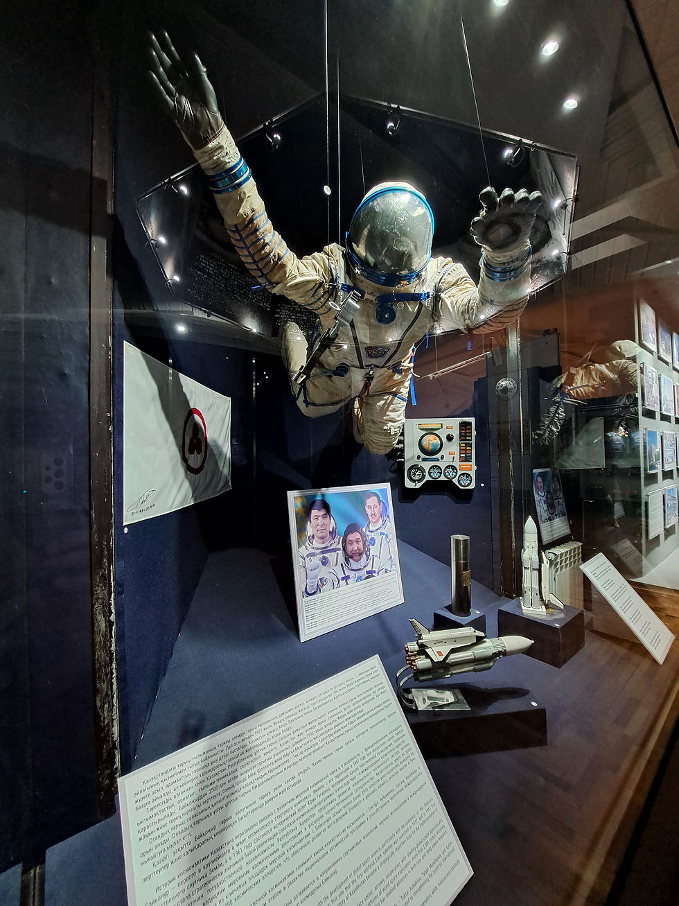 Museum display of a floating Soviet-era astronaut suit, spacecraft models, and plaques in a dark exhibit case.