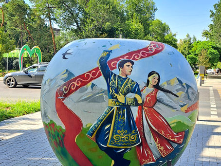 Painted apple sculpture of a dancing couple in blue and red traditional dress on a tree lined street with a clear sky.