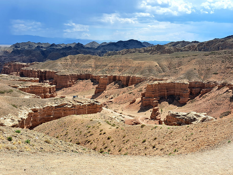 Vast red-rock canyon with layered cliffs and small hikers below a bright blue, cloud-filled sky
