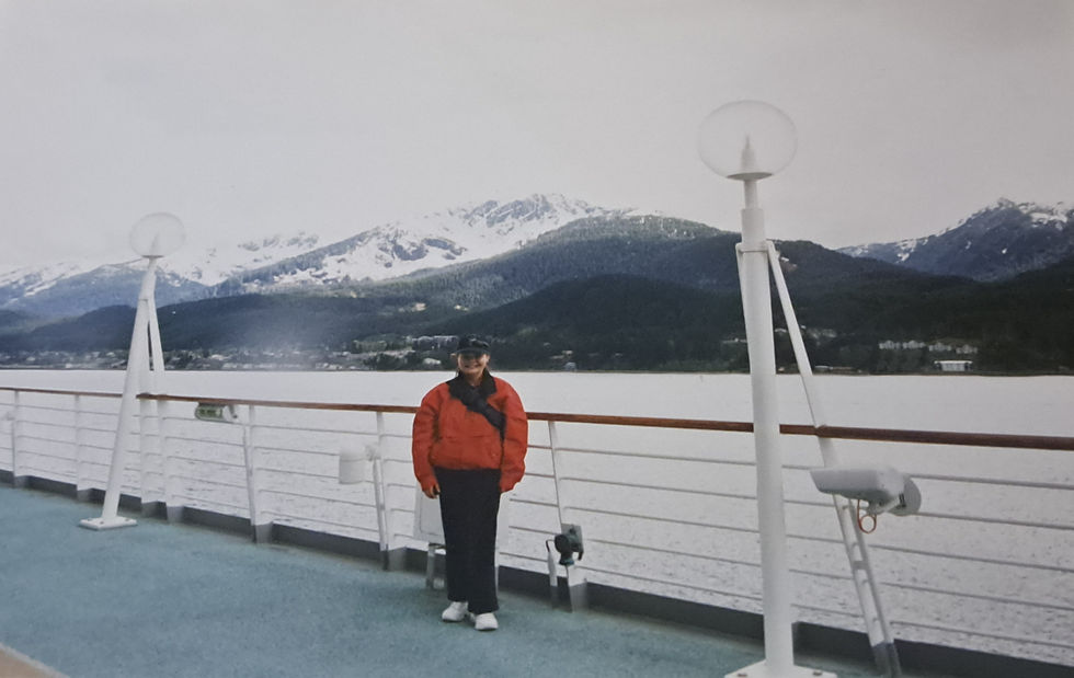 Person in a red jacket stands on a ship deck with snowy mountains and calm water in the background under a gray sky