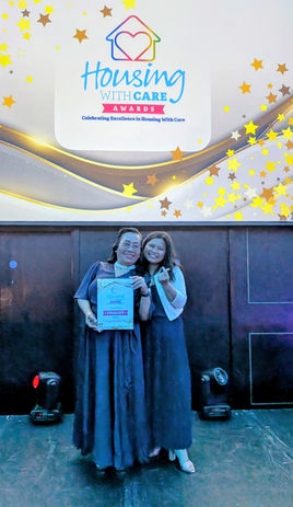 A picture of Virginia and Ruvee smiling together with the 'Housing with care' logo above them on the wall. Virginia is holding a 'finalist' certificate.