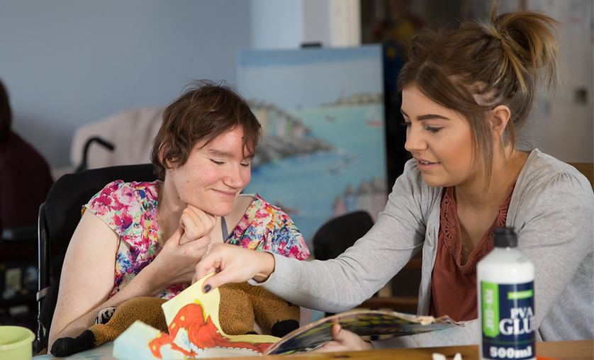 A person with disabilities and in a wheel chair with their support worker during an activity of arts and crafts with a sticker book. Both people are looking down at the book and smiling.