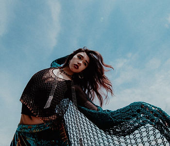 James the Seventh wearing a turquoise shrug and skirt with a black mesh top underneath against a blue sky with cirrus clouds
