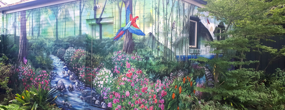 Custom exterior mural transforming a house façade into a colourful garden landscape with flowers, greenery and a painted stream, by JayMT & Silva mural artists available for projects in Germany.