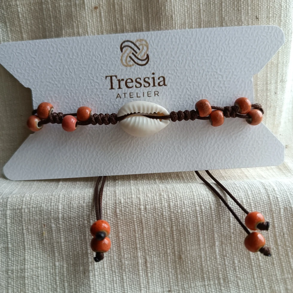 Bracelet en macramé avec coquillage cauri et perles orange présenté sur carte Tressia Atelier sur fond textile naturel