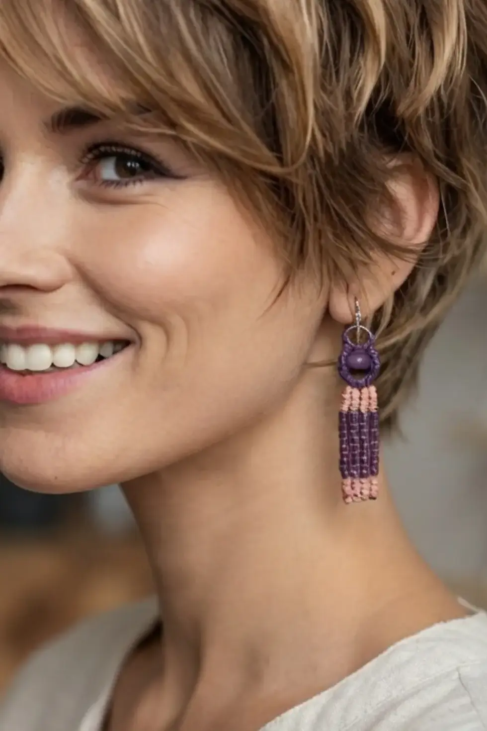 Femme aux cheveux courts portant les boucles d'oreilles pendantes en macramé violet et rose, montrant le détail des perles