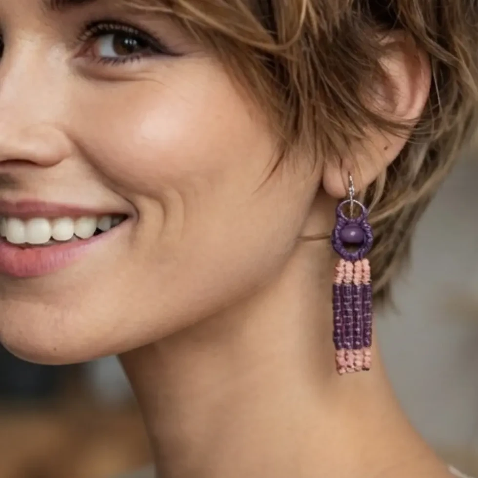Femme aux cheveux courts portant les boucles d'oreilles pendantes en macramé violet et rose, montrant le détail des perles