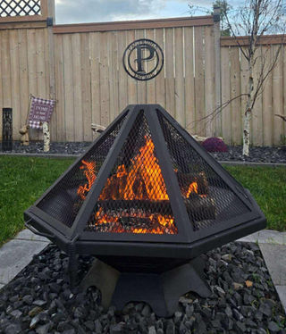 35" Octagon Fire Pit with matching Raised Spark Screen, painted black. Pictured in a yard. The fire pit is sitting on stones and surrounded by a brick border with grass around that. In the background you see a fence and border garden in front of that.