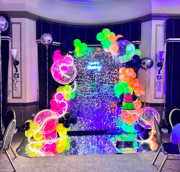 Silver shimmer wall backdrop with neon colours balloon garland, neon sign "Happy Birthday" and black lights.