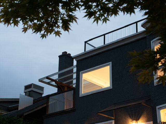 Modern house exterior at dusk with a dark facade, illuminated windows, and a rooftop railing, framed by tree branches for a cozy and contemporary look.
