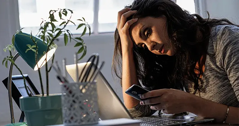 Mulher com celular na mão, em frente a um computador, com expressão frustrada.
