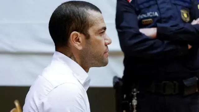 Jogador Daniel Alves, de perfil, com expressão fechada. Ao fundo, oficial de polícia de braços cruzados.