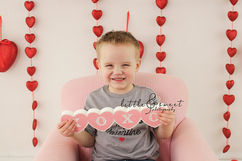 Toddler boy sitting on pink chair with red heart backdrop holding xoxo sign