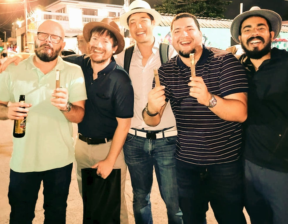 Five men smiling, holding cigars and drinks, in a lively street setting at night. Casual attire and hats, joyful mood.